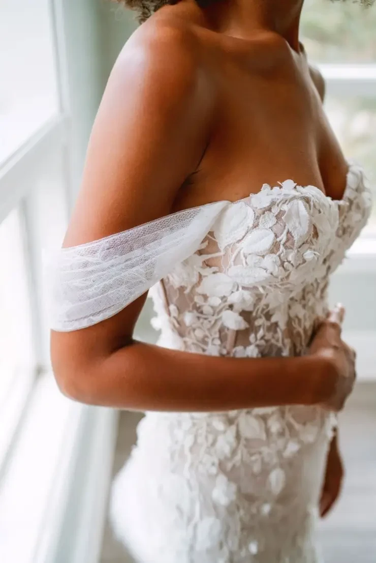 Bride in a lace-embellished, off-the-shoulder wedding gown stands by a sunlit window. The elegant dress features floral patterns, conveying romance.