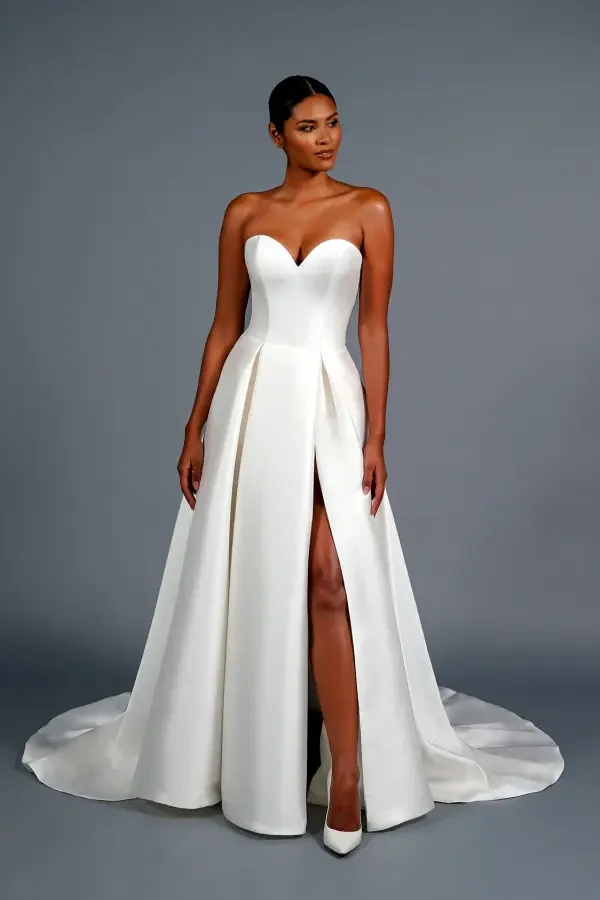Elegant woman in a strapless white gown with a sweetheart neckline and high slit, exuding confidence, standing against a gray background.