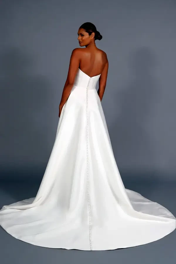A woman stands in a strapless white wedding gown with a long train and button details. She faces away against a gray background, conveying elegance.