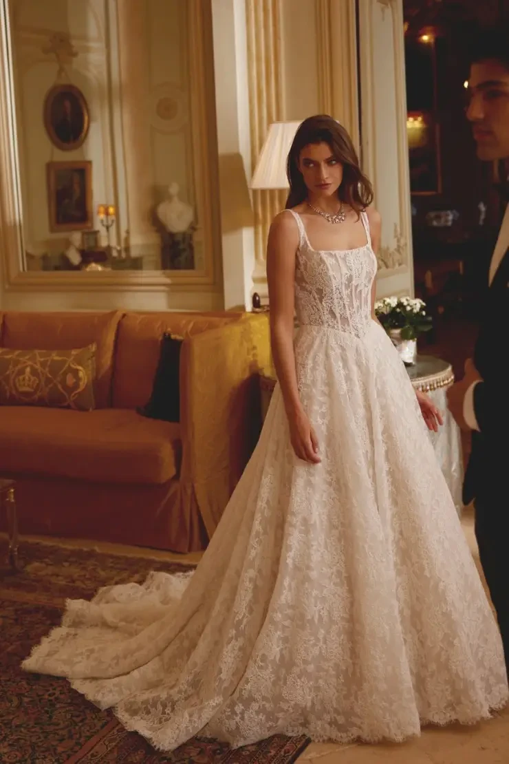 A woman stands in an elegant room wearing a beautiful lace wedding dress with a flowing train. The interior features soft lighting, luxurious furnishings, and decorative elements.