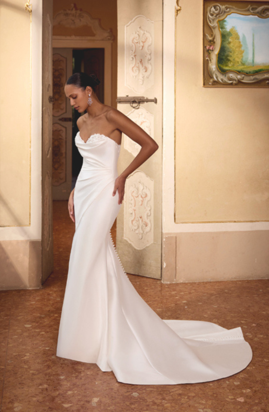 Bride in elegant, strapless white gown with long train, stands gracefully in ornate room with soft lighting. A framed landscape painting hangs on the wall.