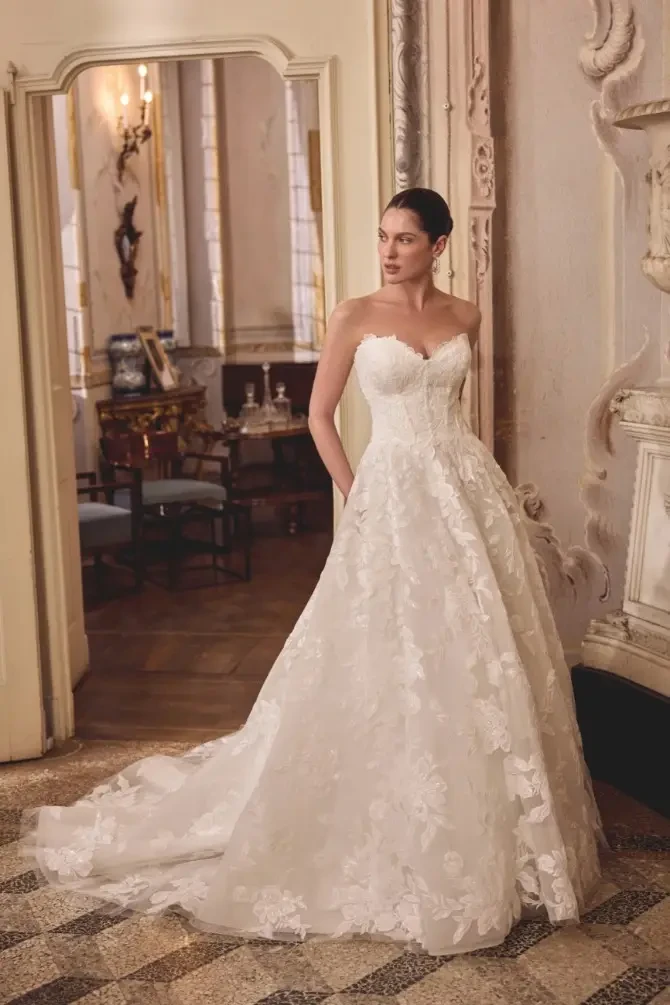 A woman in a strapless white wedding gown with floral details stands elegantly in a beautifully decorated room.