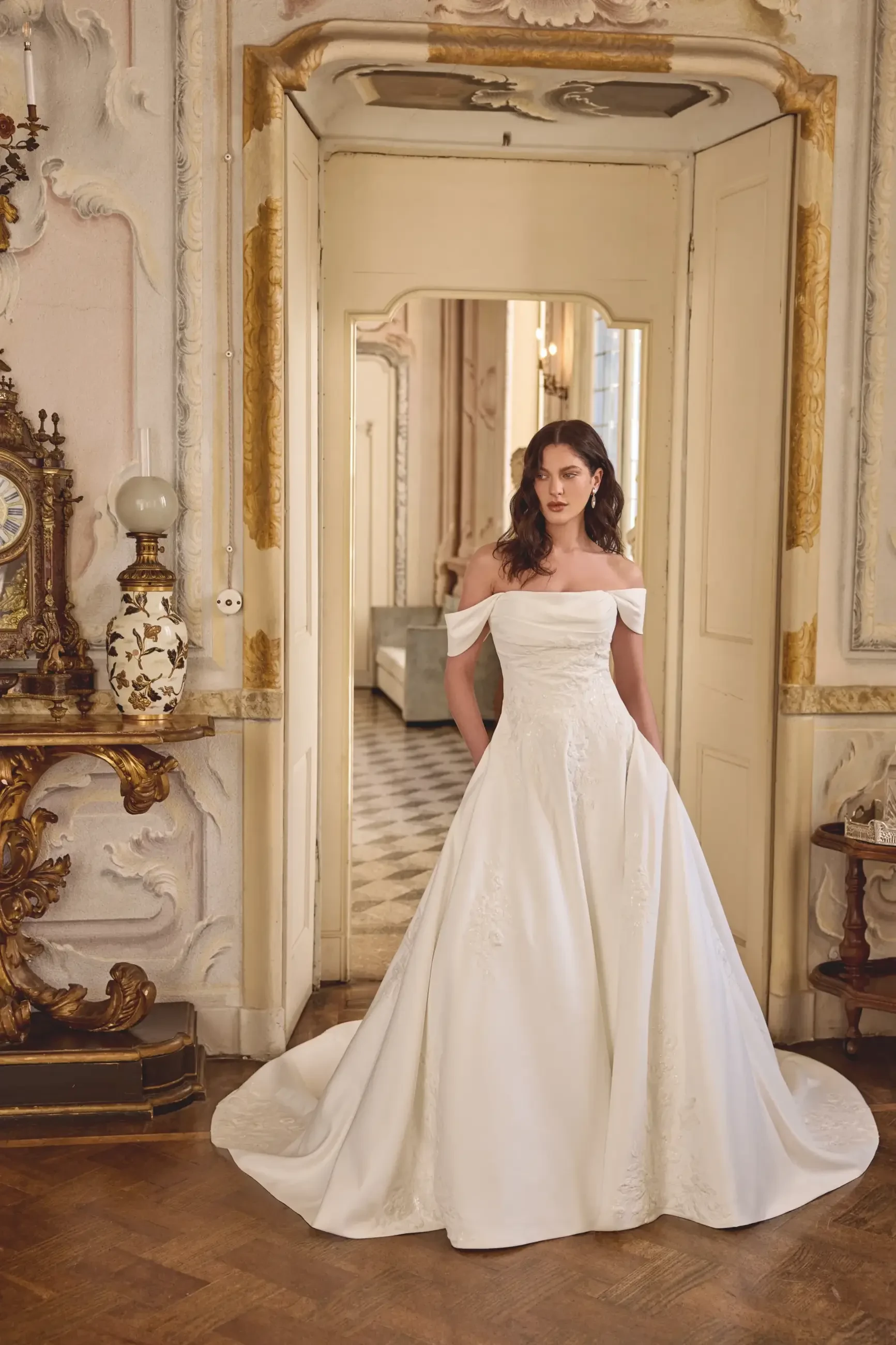 A woman wearing an off-the-shoulder white wedding dress stands in a beautifully decorated room with ornate furniture and mirrors.