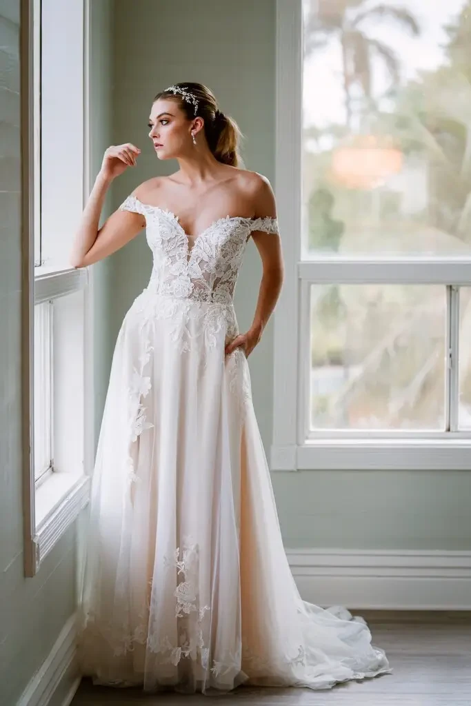 A bride in an off-shoulder lace wedding dress stands by a window, gazing outside. Soft light highlights the delicate floral patterns on the gown.