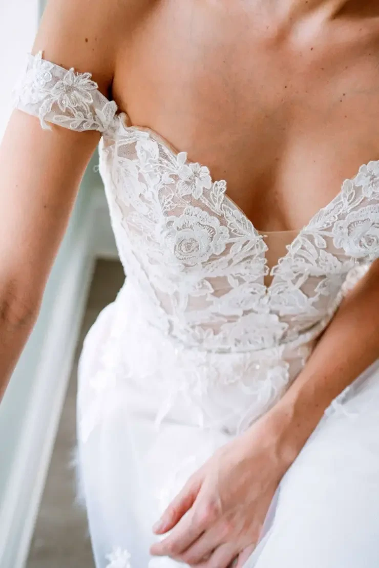 Close-up of a bride in an off-the-shoulder lace wedding dress, showcasing intricate floral patterns. The mood is elegant and romantic.