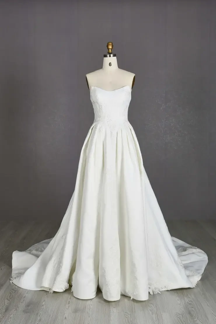 A simple, elegant white wedding dress displayed on a mannequin. The dress has a strapless design and a flowing skirt that extends into a train.