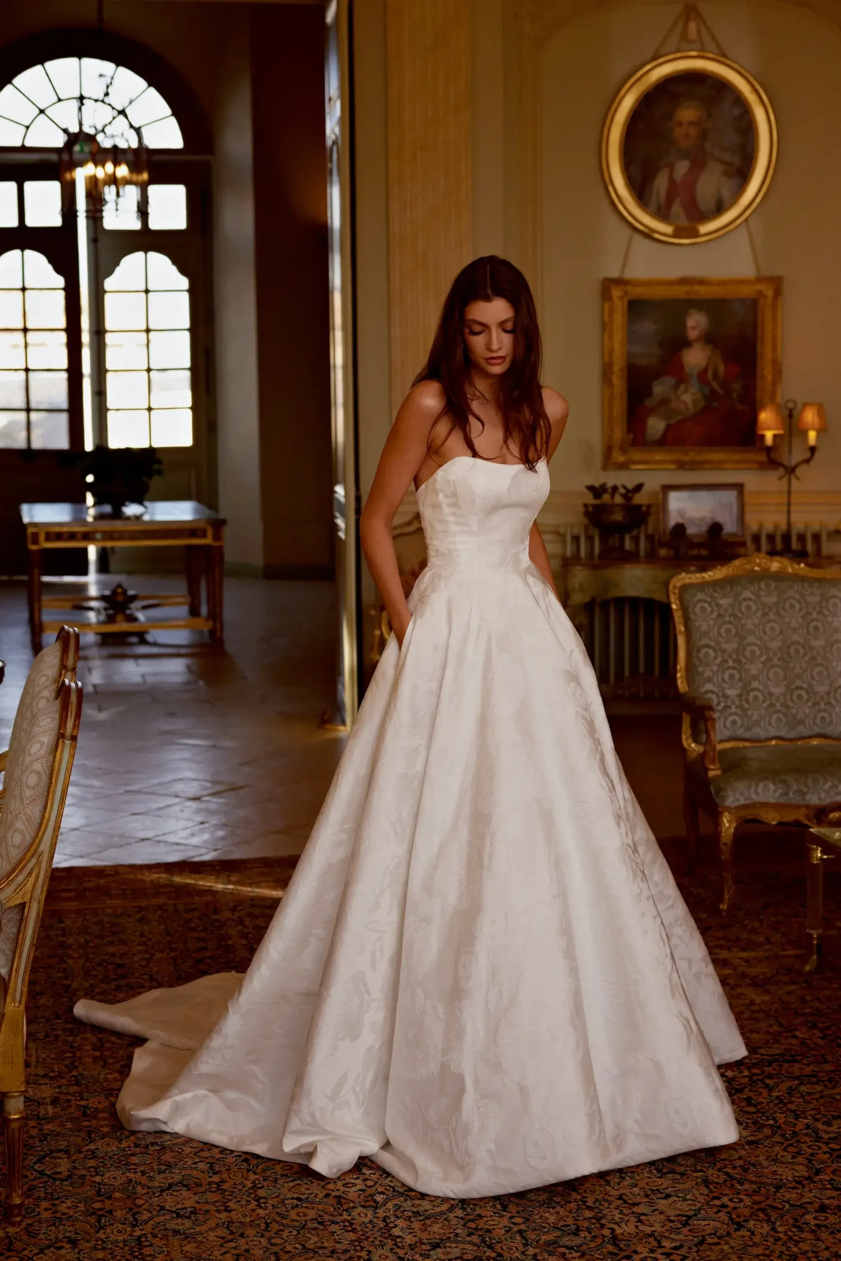 A woman in a strapless white wedding gown stands in an ornate room with vintage furniture, soft lighting, and large windows, exuding elegance and serenity.