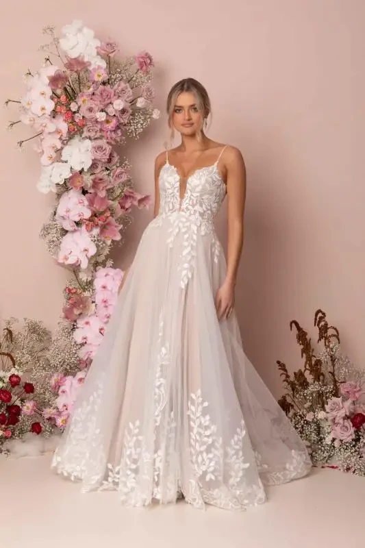 A woman in an elegant, white floral lace gown stands before a pastel pink background with a lush floral arrangement of pink and white roses.