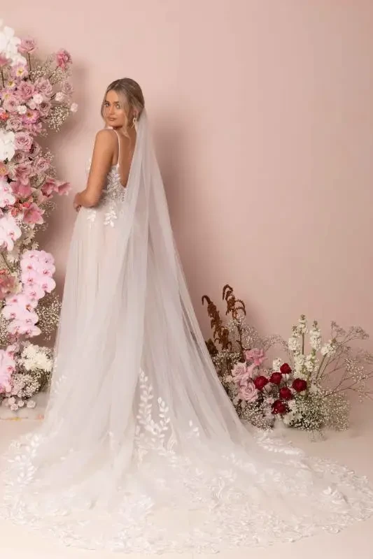 Bride in an elegant lace wedding dress and long veil stands amidst pink and white flowers against a soft pink backdrop, exuding romance and grace.
