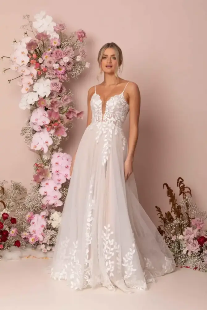 A woman in an elegant white bridal gown with lace patterns stands beside a vibrant floral arrangement of pink and white flowers, set against a soft pink background.