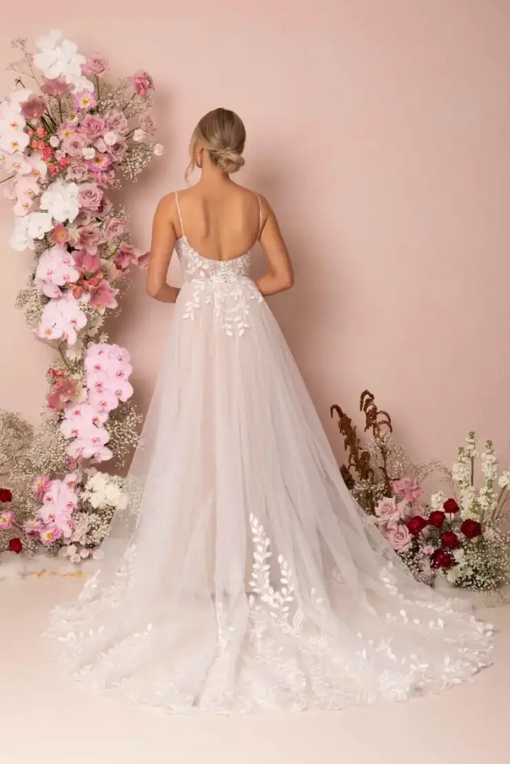 A woman in an elegant, backless white wedding dress stands facing a pastel pink wall. The floor-length gown has delicate floral embroidery, and she is surrounded by vibrant pink and white floral arrangements, creating a romantic atmosphere.