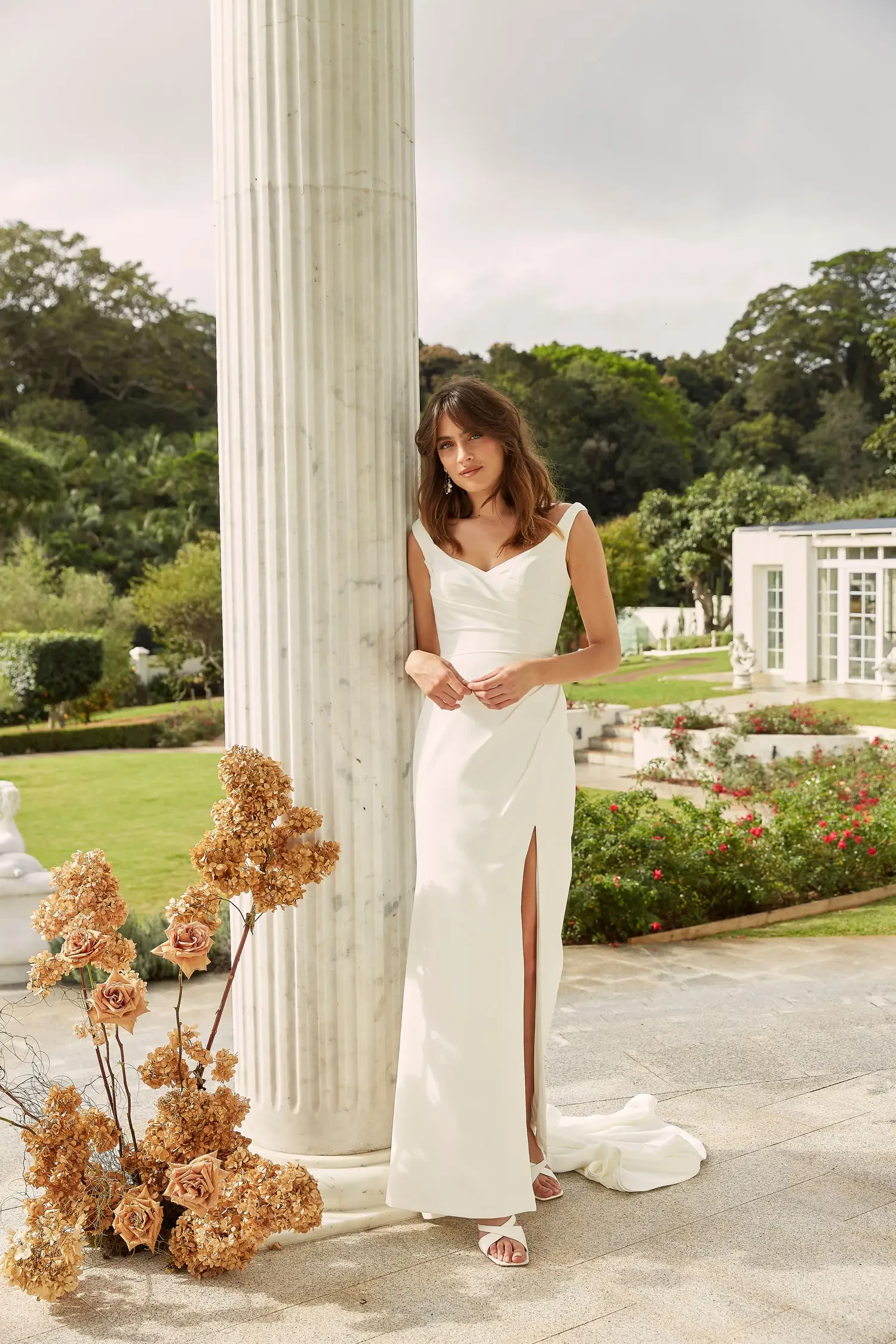 A woman in an elegant white gown with a thigh-high slit leans against a tall column in a lush garden. The background includes manicured lawns and a classical building, evoking a serene and timeless mood.