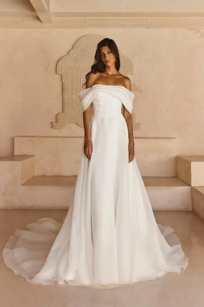 A model wearing a long, elegant white wedding dress with off-the-shoulder sleeves, standing gracefully in a minimalist setting.