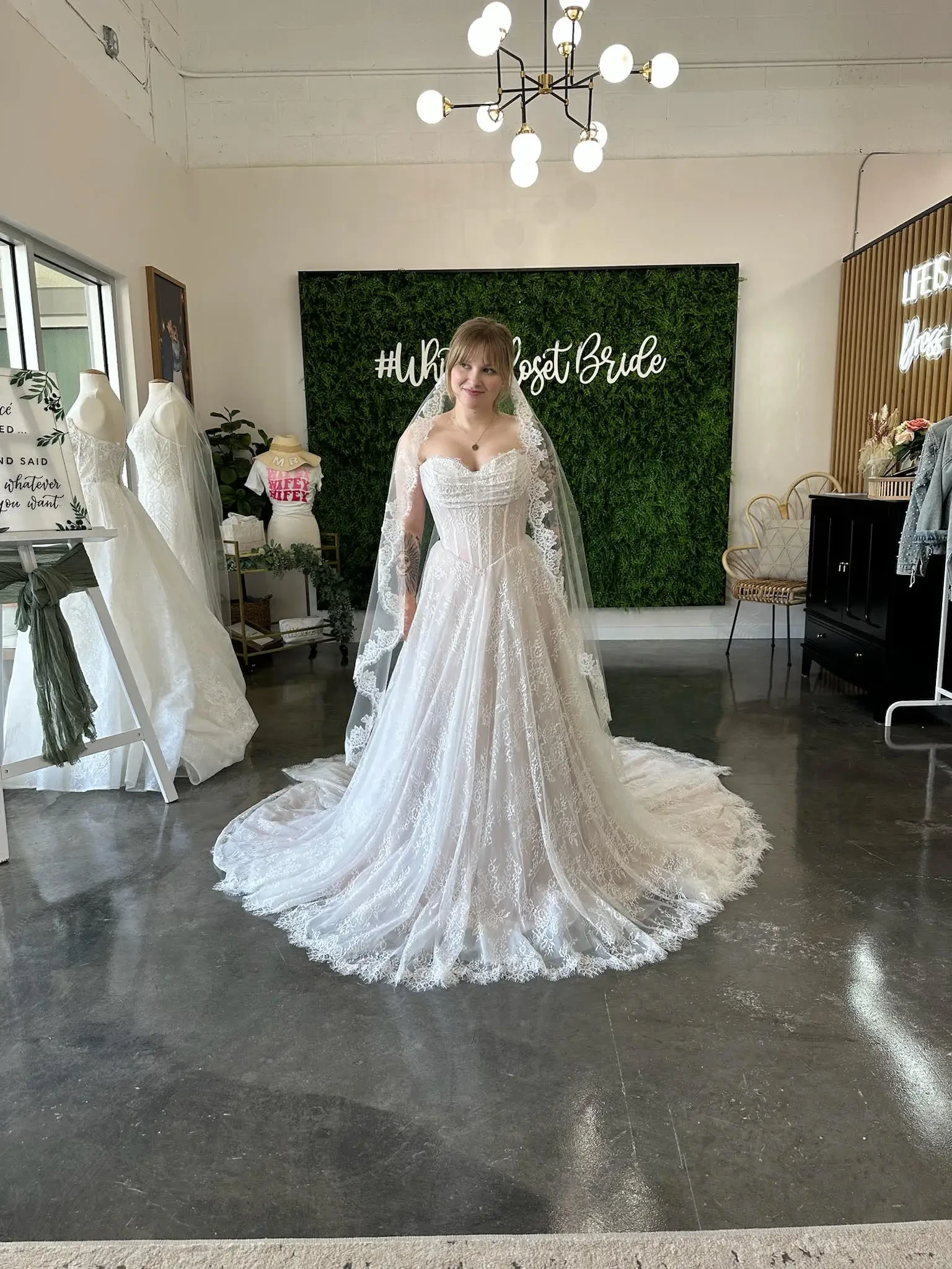 A bride stands joyfully in a lace wedding gown and veil, with a sign 