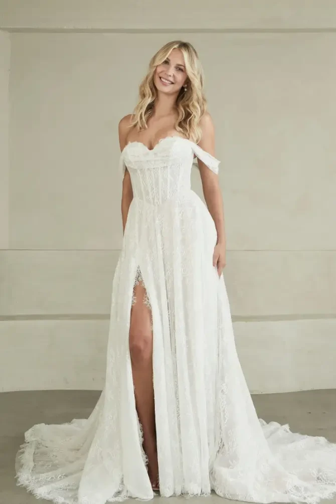 A smiling woman wearing a flowing white wedding dress with a thigh-high slit, standing in a minimalist room.