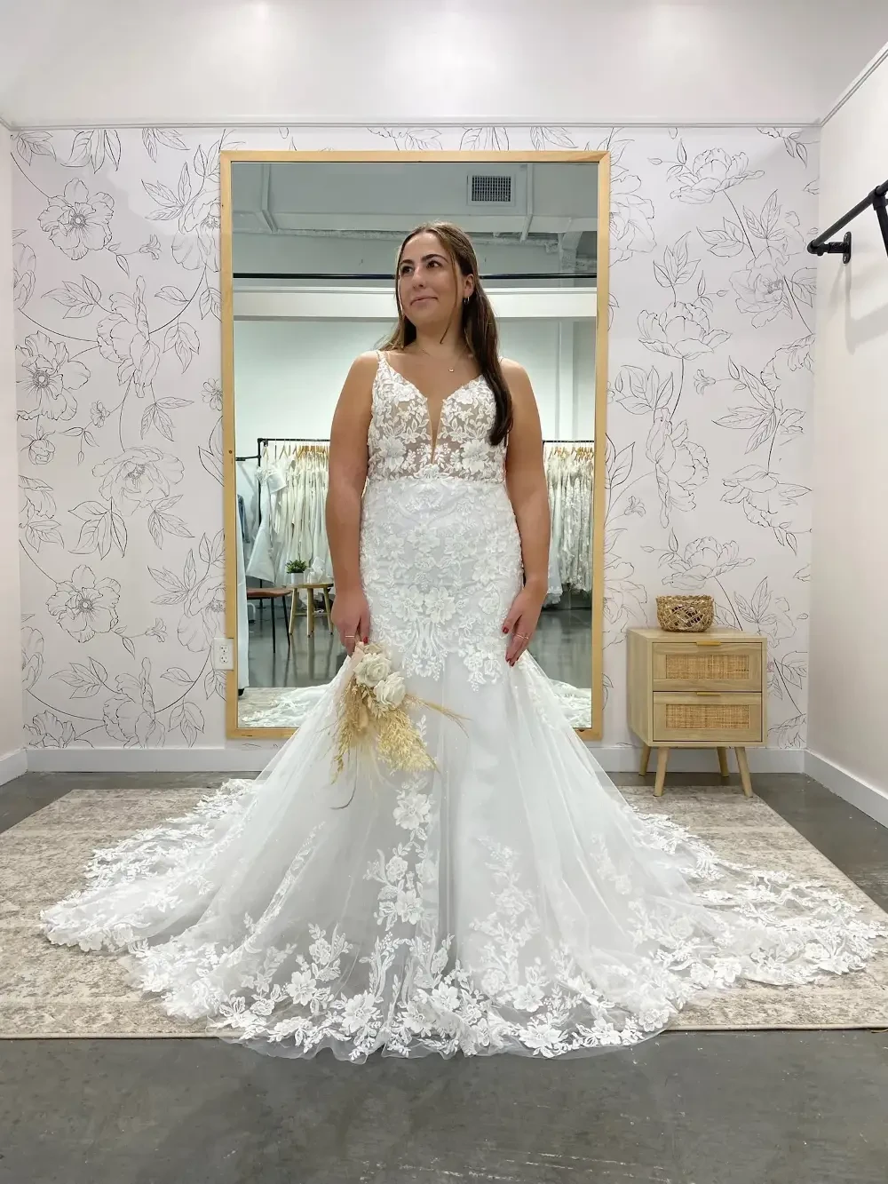 A woman wearing a white lace wedding gown stands in front of a mirror, holding a bouquet, in a bridal shop setting.