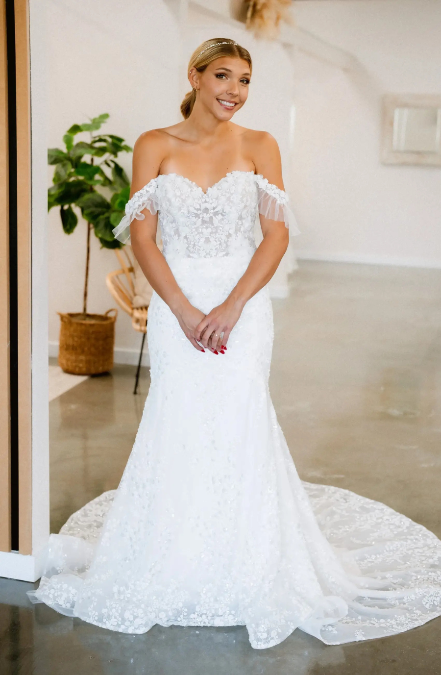 A smiling bride stands in a sleek room wearing an off-shoulder, lace wedding gown with a long train. A potted plant and wicker chair are in the background.