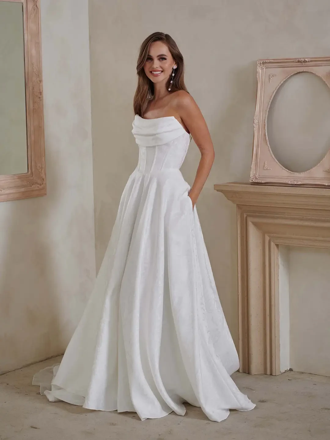 A woman in a strapless white wedding dress stands smiling in an elegant room. She poses near an ornate mirror and mantel, exuding a joyful, serene vibe.