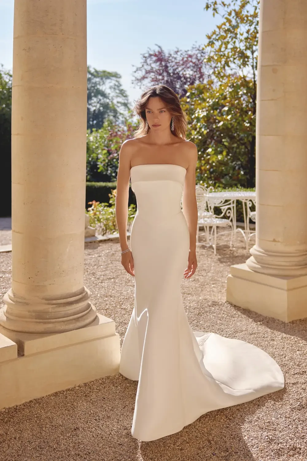 A woman in a strapless white dress standing outdoors near columns and trees.