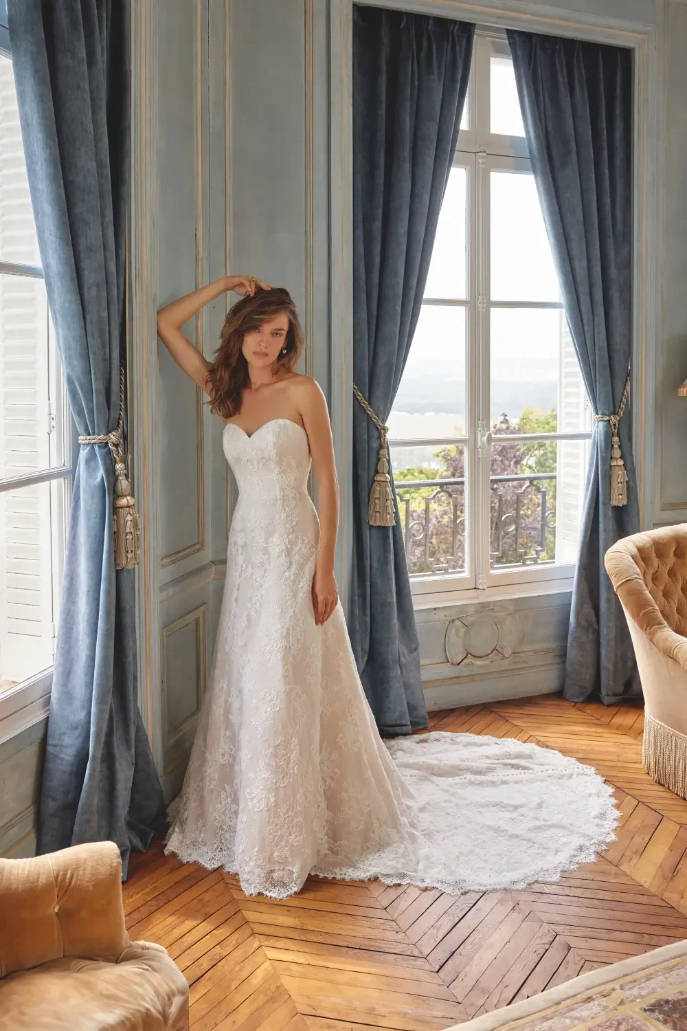 A woman in a strapless white wedding dress stands by a window in a room with blue drapes and wooden flooring.