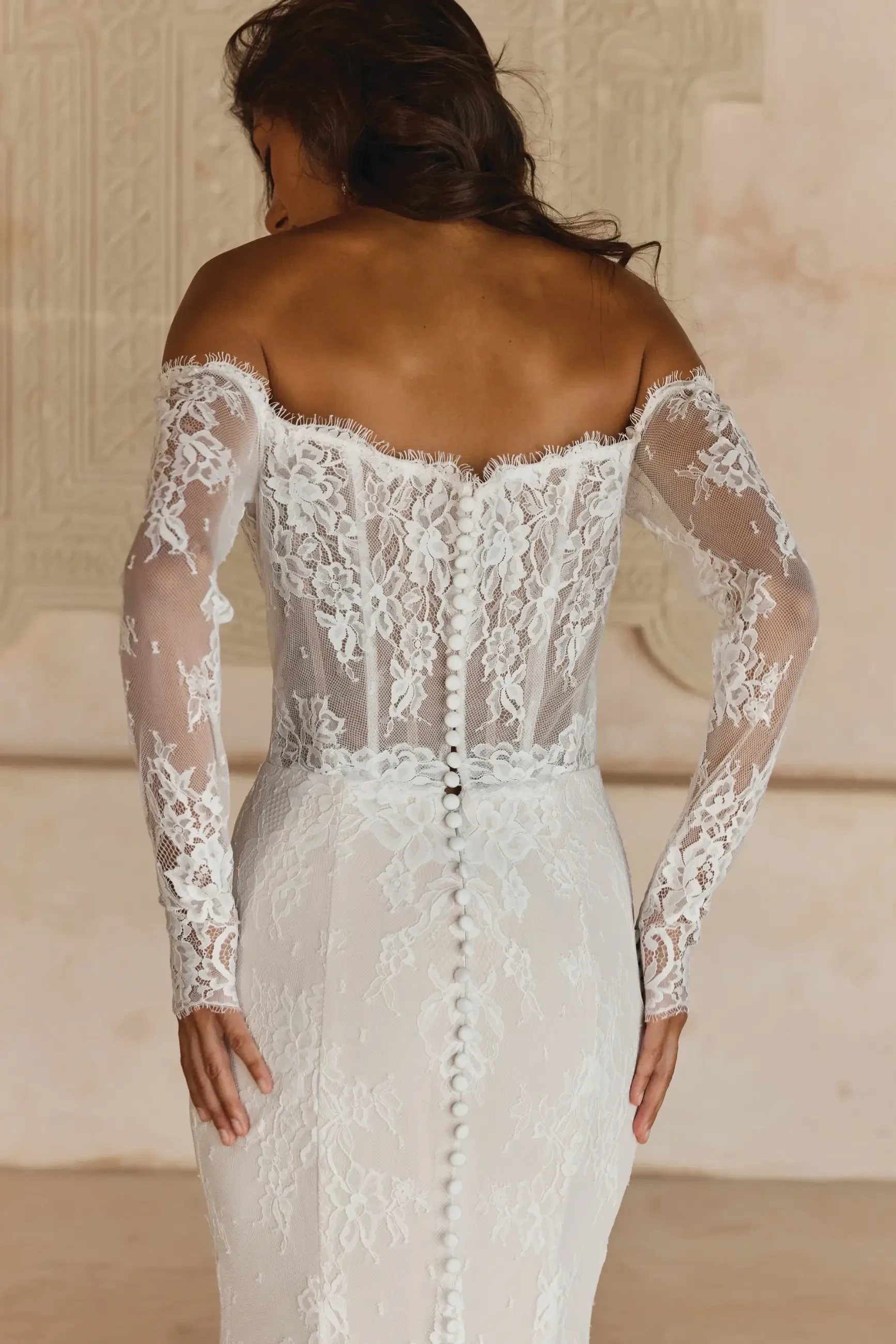 Back view of a woman in an elegant, off-the-shoulder lace wedding dress with delicate floral patterns and buttons. The setting is softly lit and ornate.