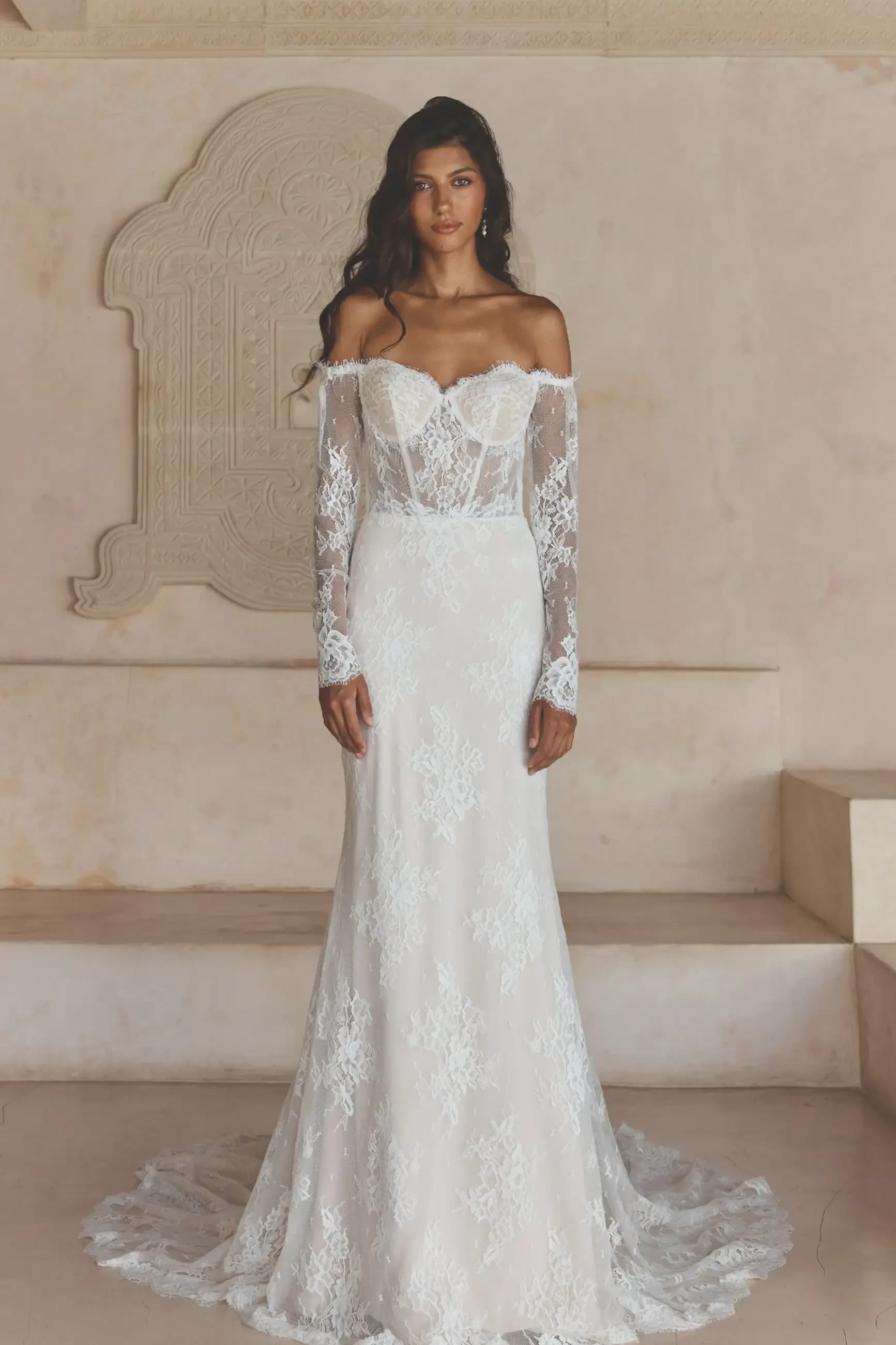 A woman in an elegant, off-the-shoulder lace wedding gown stands before a textured stone wall. The dress features a fitted bodice and a flowing train.