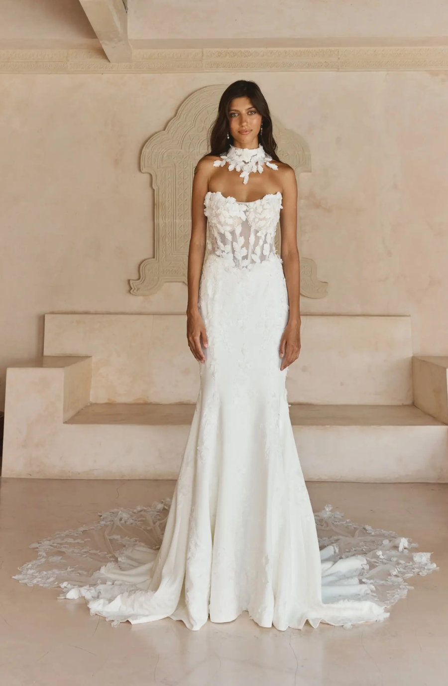 Bride in an elegant, strapless lace wedding gown stands on a polished floor. The dress features intricate floral detailing and a long train. The setting is minimalist with a beige backdrop, conveying a serene and sophisticated tone.