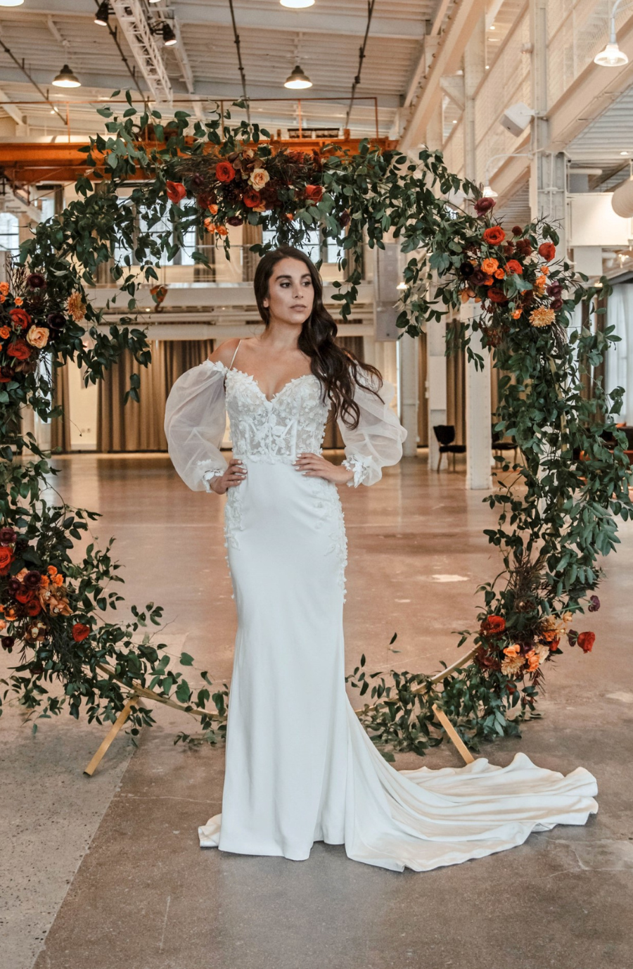 A woman in a sleek white wedding dress with sheer puffy sleeves stands confidently in front of a large, circular floral arch, featuring lush green leaves and vibrant orange flowers. The industrial hall setting is softly lit, adding an elegant, modern ambiance.