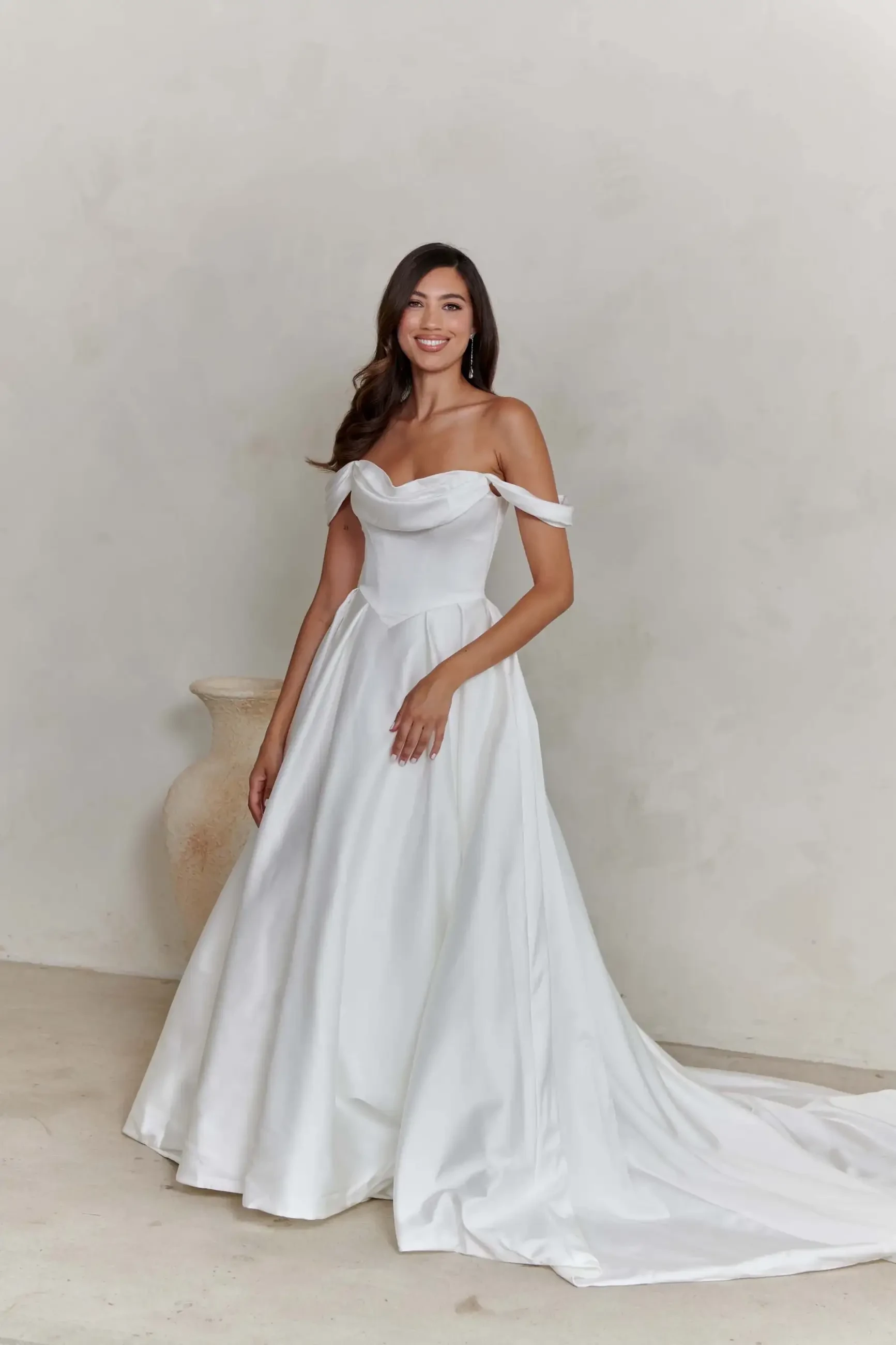 A woman in a white off-shoulder wedding dress stands smiling. She is in a minimalistic setting with a light-colored background and a vase nearby.